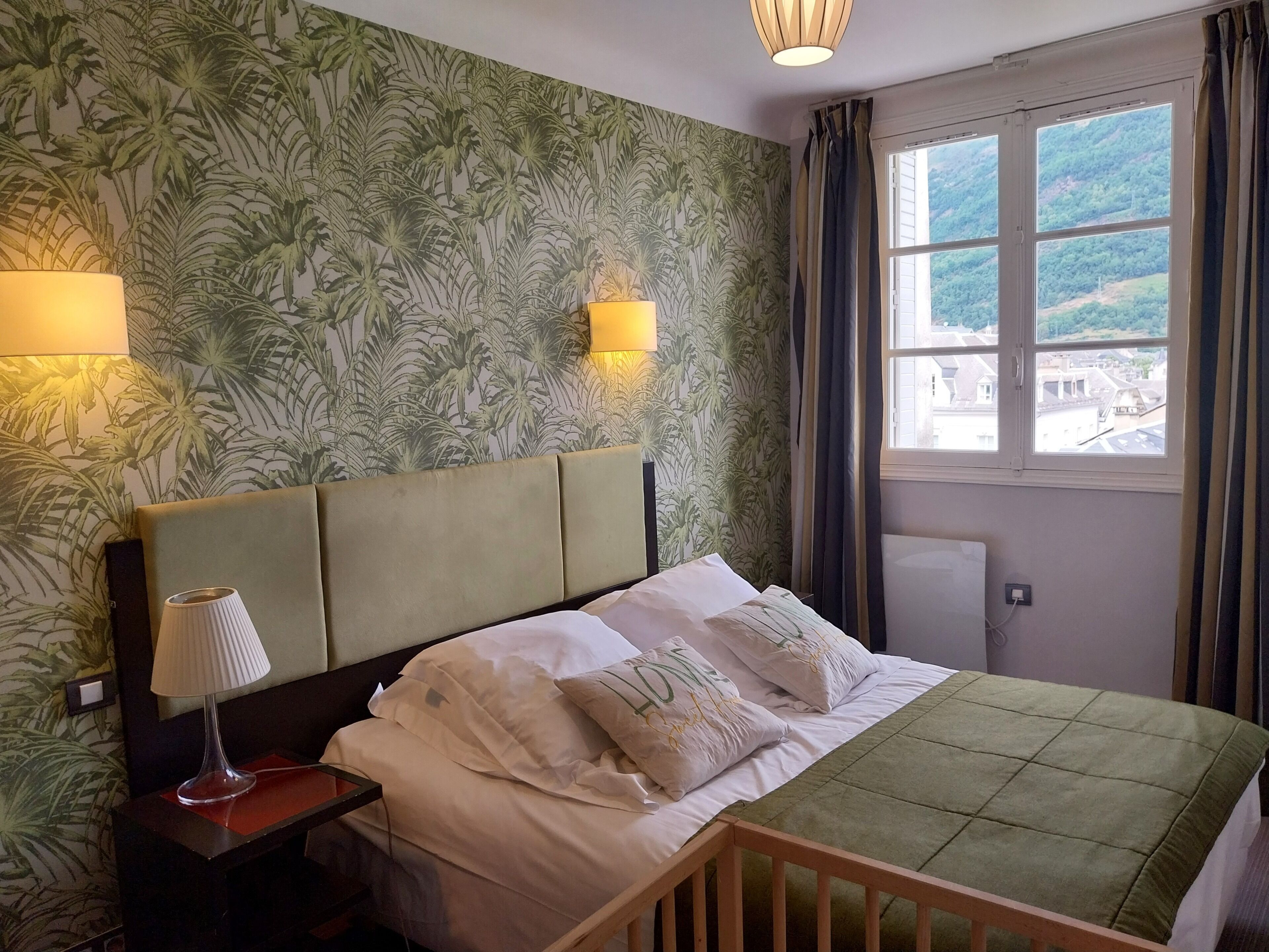 Een hotelkamer met een bed, twee bedlampen, een raam met gordijnen en een muur met tropisch behang.