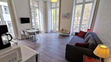Uma sala de estar moderna com sofá, máquina de café, mesa de jantar com cadeiras e grandes janelas com painéis decorativos.