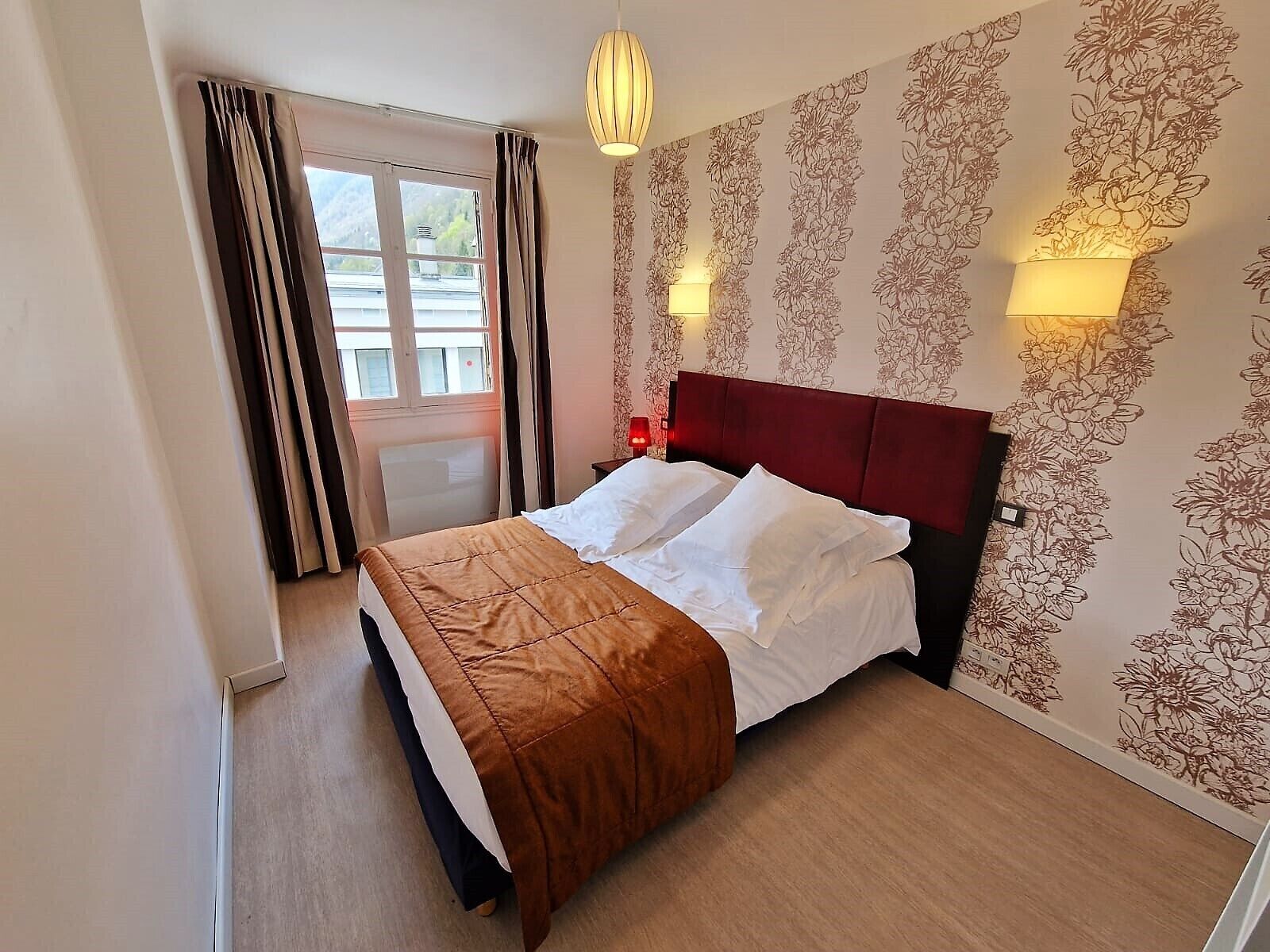 Hotelkamer met een bed, een raam met gordijnen, een wand met bloemenbehang en twee wandlampen.