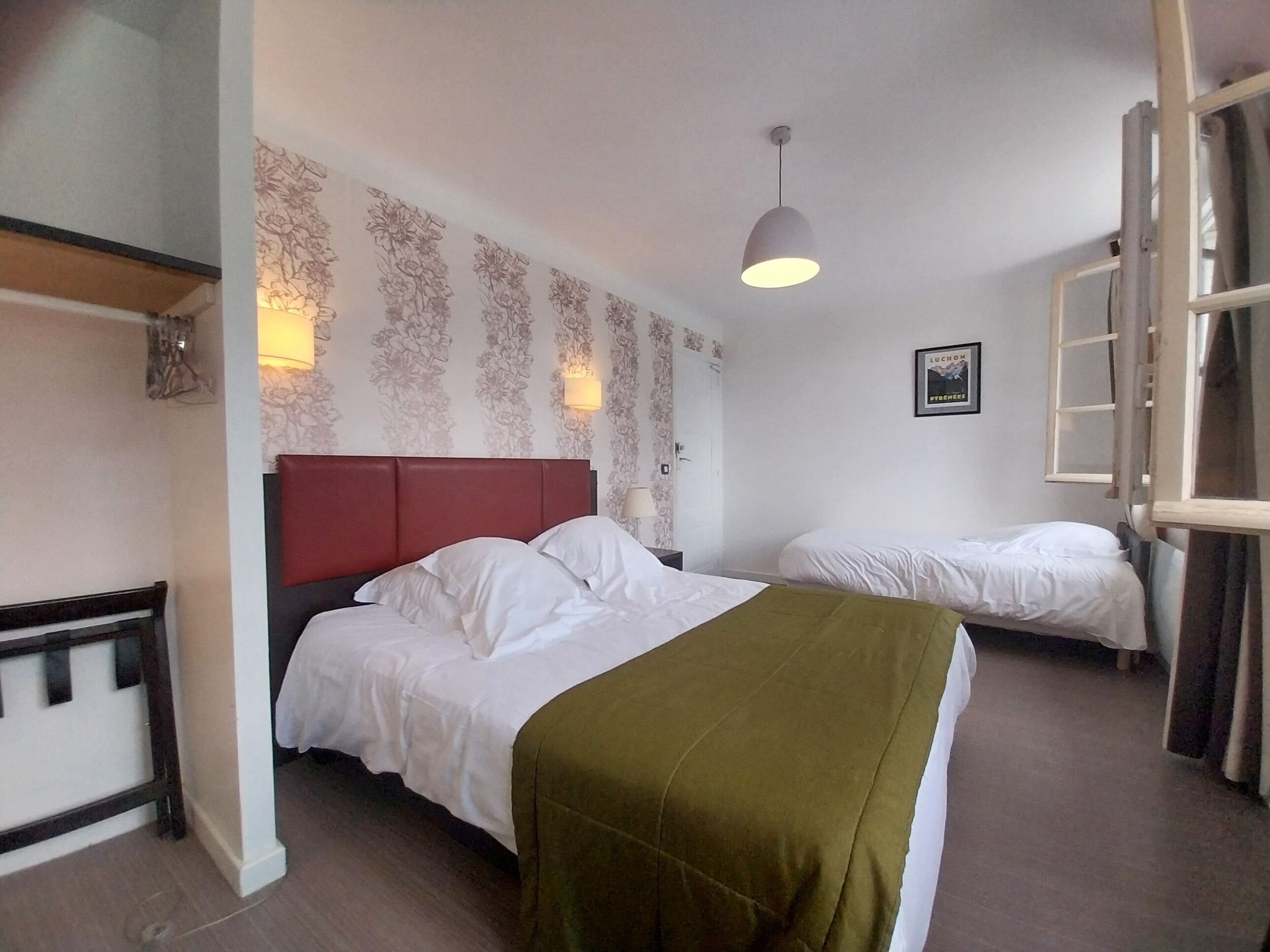 Habitación de hotel con dos camas, cabecera roja, luz empotrada en la pared y ventana con cortinas.