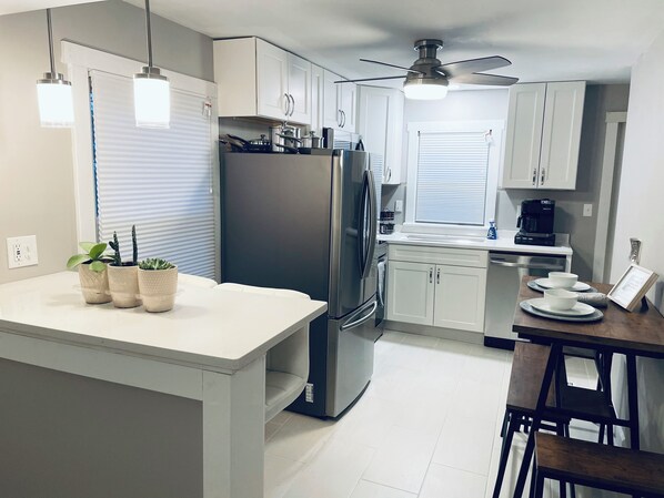 A modern kitchen with white cabinets, a stainless steel refrigerator, and a built-in oven. There are three potted plants on the counter and a ceiling fan above.