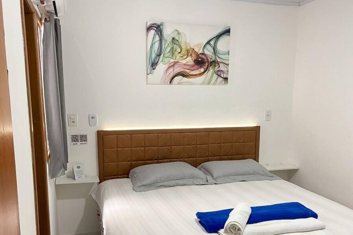 Een hotelkamer met een bed, hoofdbord, een wandairconditioner, nachtkastje en een schilderij.