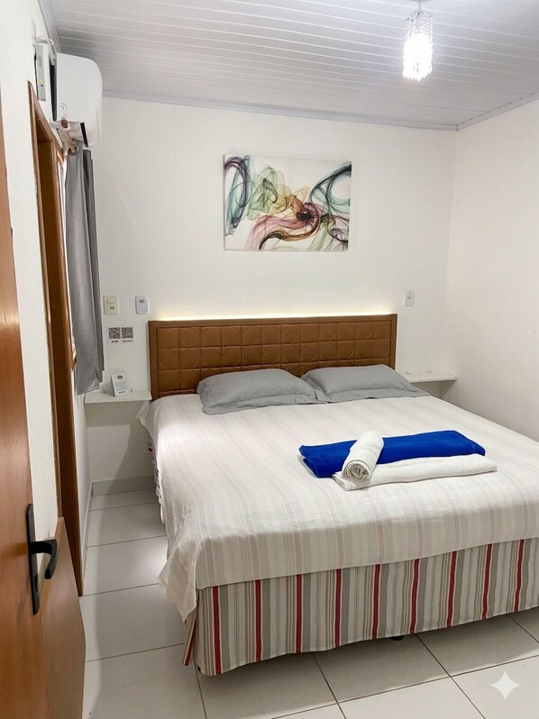 Une chambre d’hôtel comprenant un lit, une tête de lit, un climatiseur fixé au mur, une table de chevet et une commode, ainsi qu’un tableau.