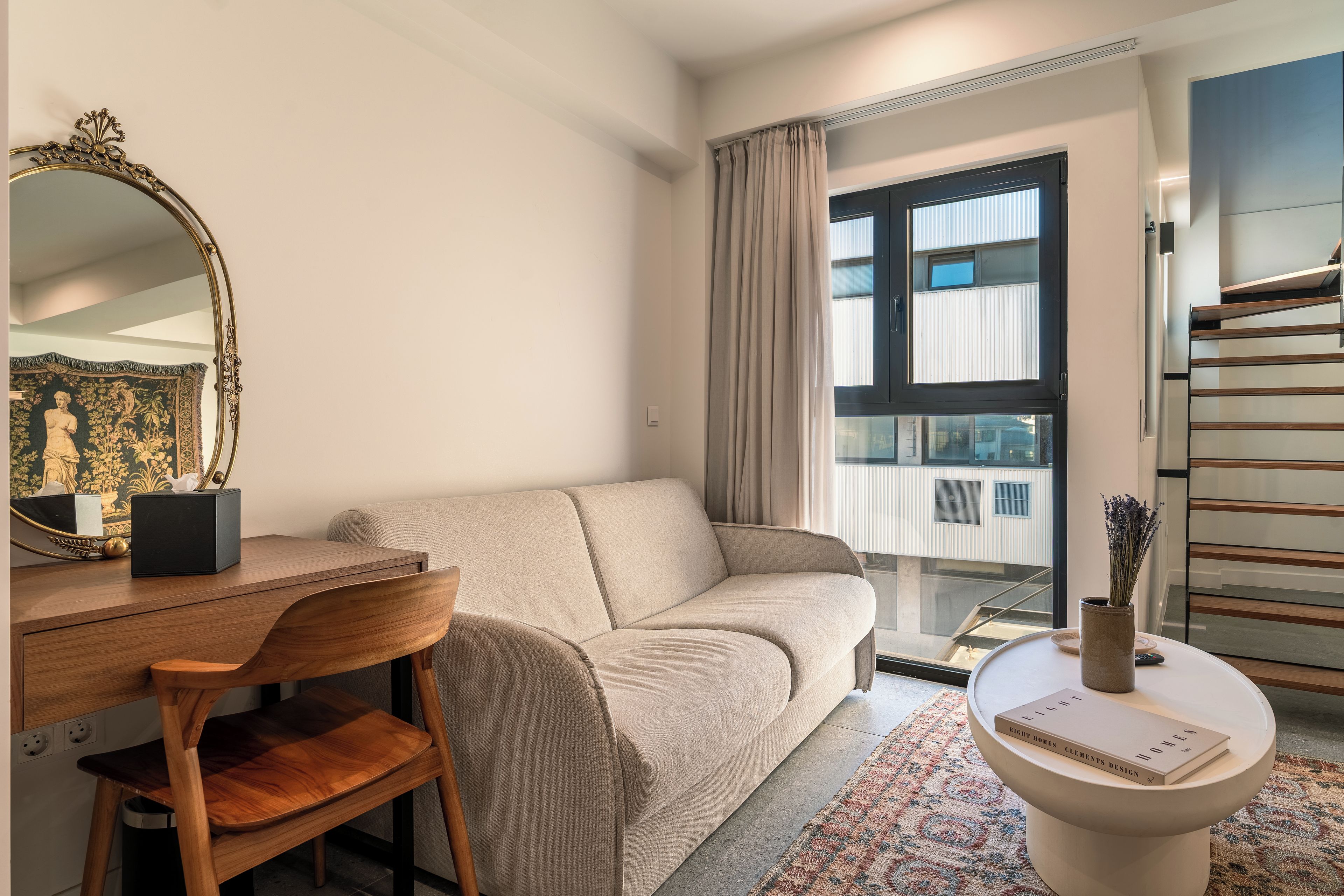Una sala moderna con un sofá, una silla de madera, una mesa redonda de centro, un espejo decorativo y una ventana con cortinas.