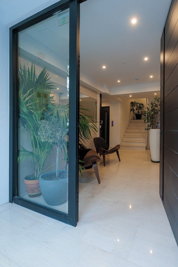 A modern hallway with a glass door, potted plants, a chair, and a staircase leading to an upper level. - Fos (Athens)