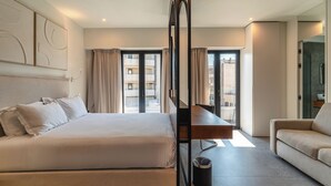 A modern hotel room with a bed, a sofa, a dining table, and a view of the cityscape. - Fos (Athens)
