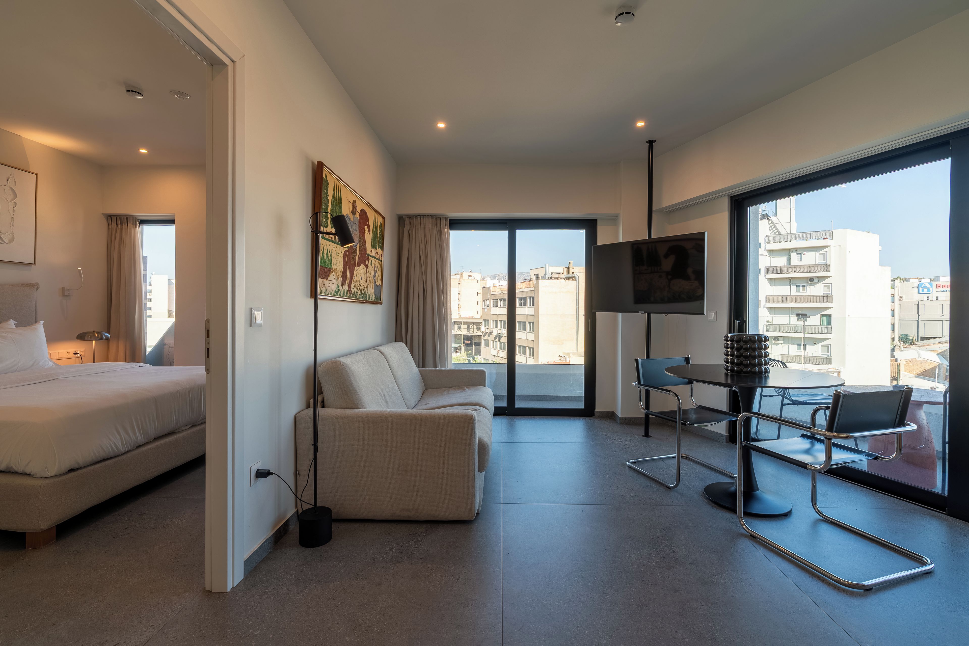 A modern hotel room with a bed, a sofa, a dining table with chairs, a TV, and a large window offering a city view.