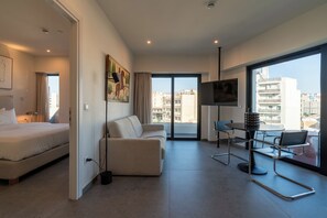 A modern hotel room with a bed, a sofa, a dining table with chairs, a TV, and a large window offering a city view. - Fos (Athens)
