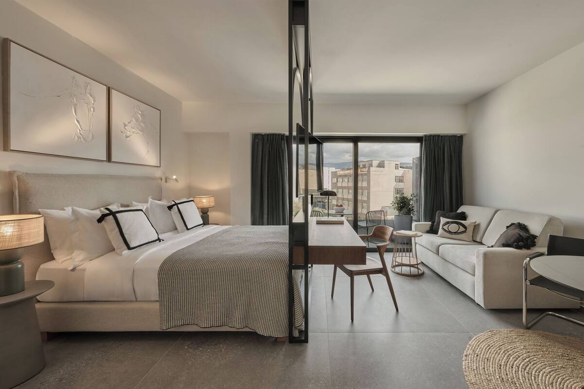 A modern hotel room with a bed, a sofa, a dining table, and a view of the city through large windows.