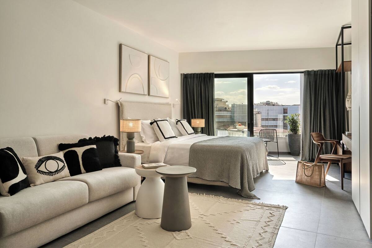 A modern hotel room with a bed, a sofa, a small table, and a balcony with a view of the city.
