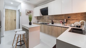 Une cuisine moderne avec des meubles blancs, un pare-feu en bois et une cuisinière encastrée. On y trouve deux tabourets blancs et un petit îlot central avec un évier.