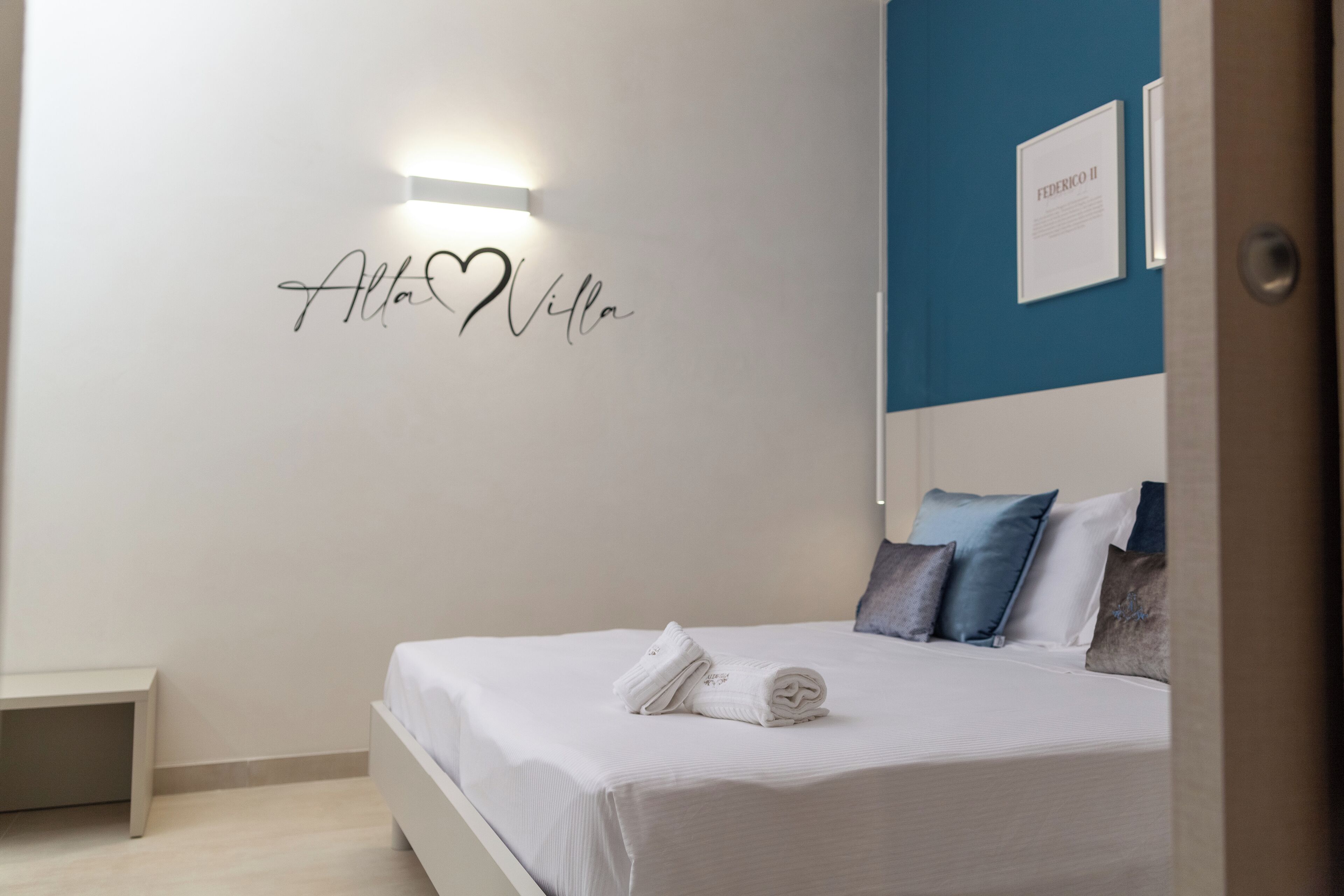 Una cama bien tendida con sábanas blancas y una toalla enrollada, una pared con un cartel con un nombre y un certificado enmarcado.