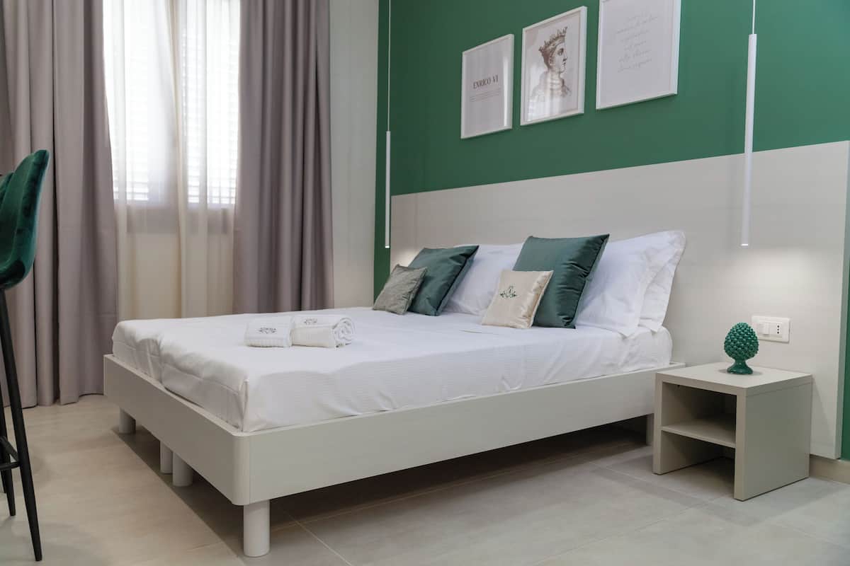 A neatly made bed with white bedding and multiple pillows, a small bedside table with a green decorative item, and a green wall with framed pictures.