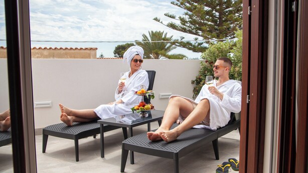 Dos personas descansando en sillones de exterior con una mesa entre ellas, sosteniendo bebidas y un cuenco de fruta.