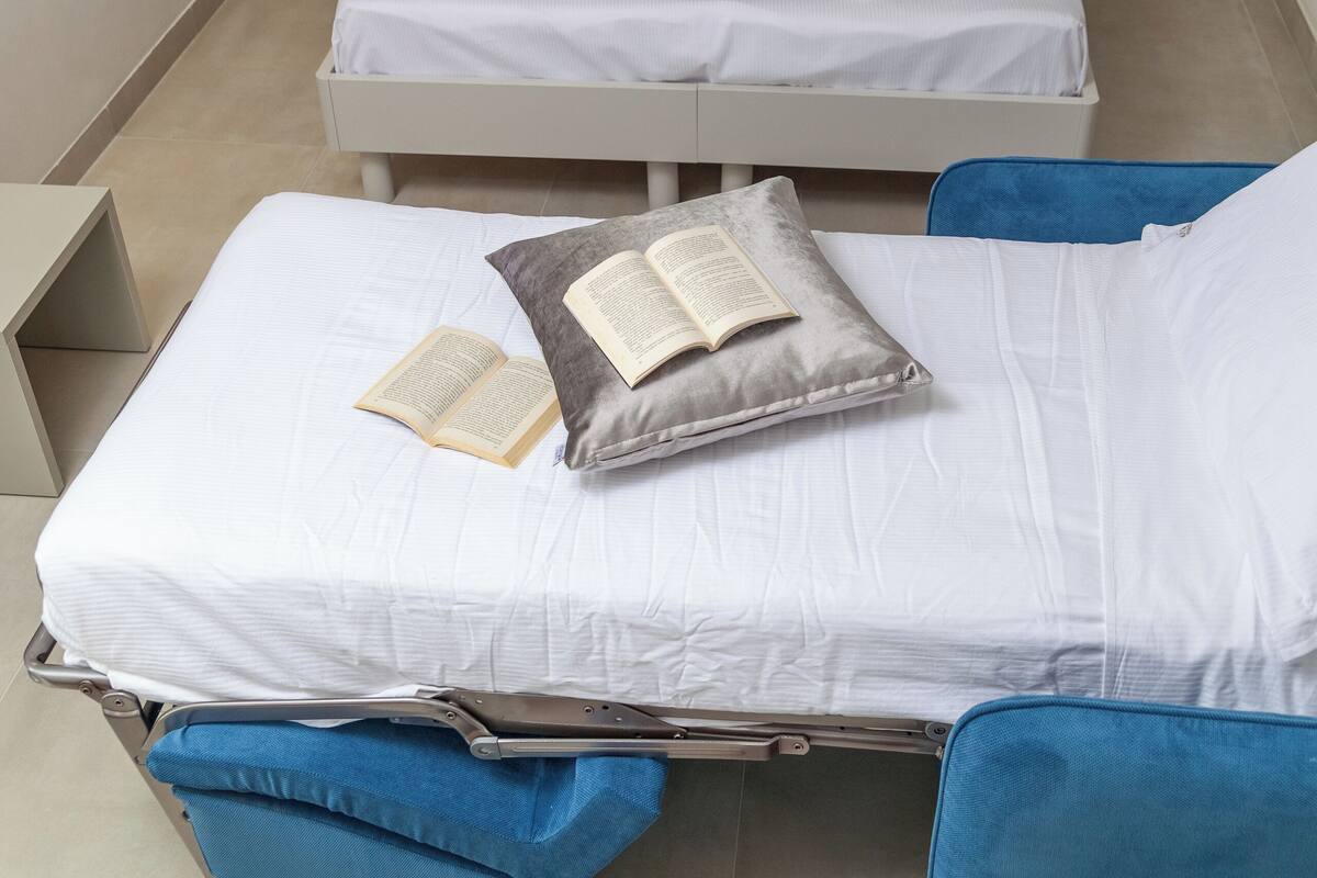 Two beds with white bedding and blue pillows, a bedside table with a book, and a blue chair.
