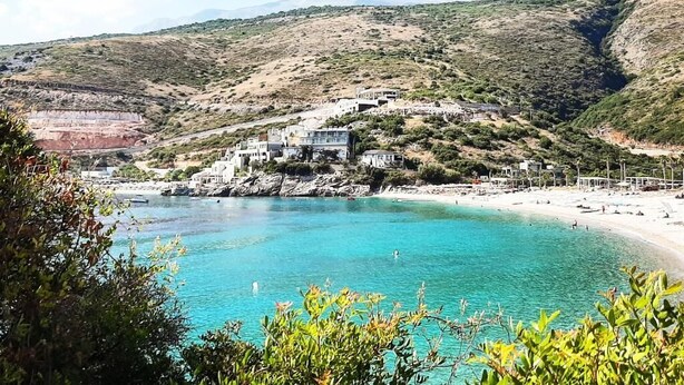 A coastal area with a beach, clear blue waters, and a hillside with buildings. - Social Camping JAL (Himarë)