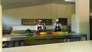 A kitchen with two chefs preparing food, a stainless steel counter, and shelves with kitchenware. - Social Camping JAL (Himarë)