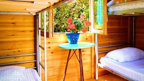 A wooden cabin room with a bunk bed, a small round table with a blue pot of flowers, and a window with a view of greenery. - Social Camping JAL (Himarë)