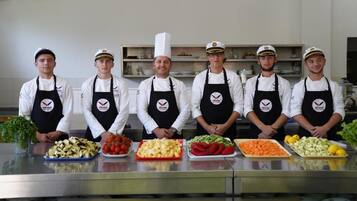 Six chefs in white shirts and black aprons with a logo, standing in a kitchen with various vegetables on the counter.