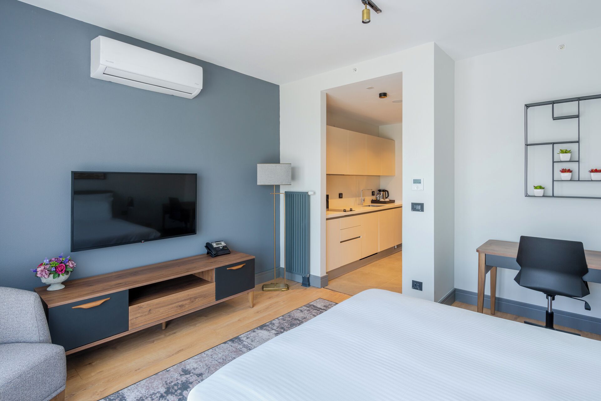 A modern hotel room with a large bed, a TV, a desk, and a kitchenette in the background.