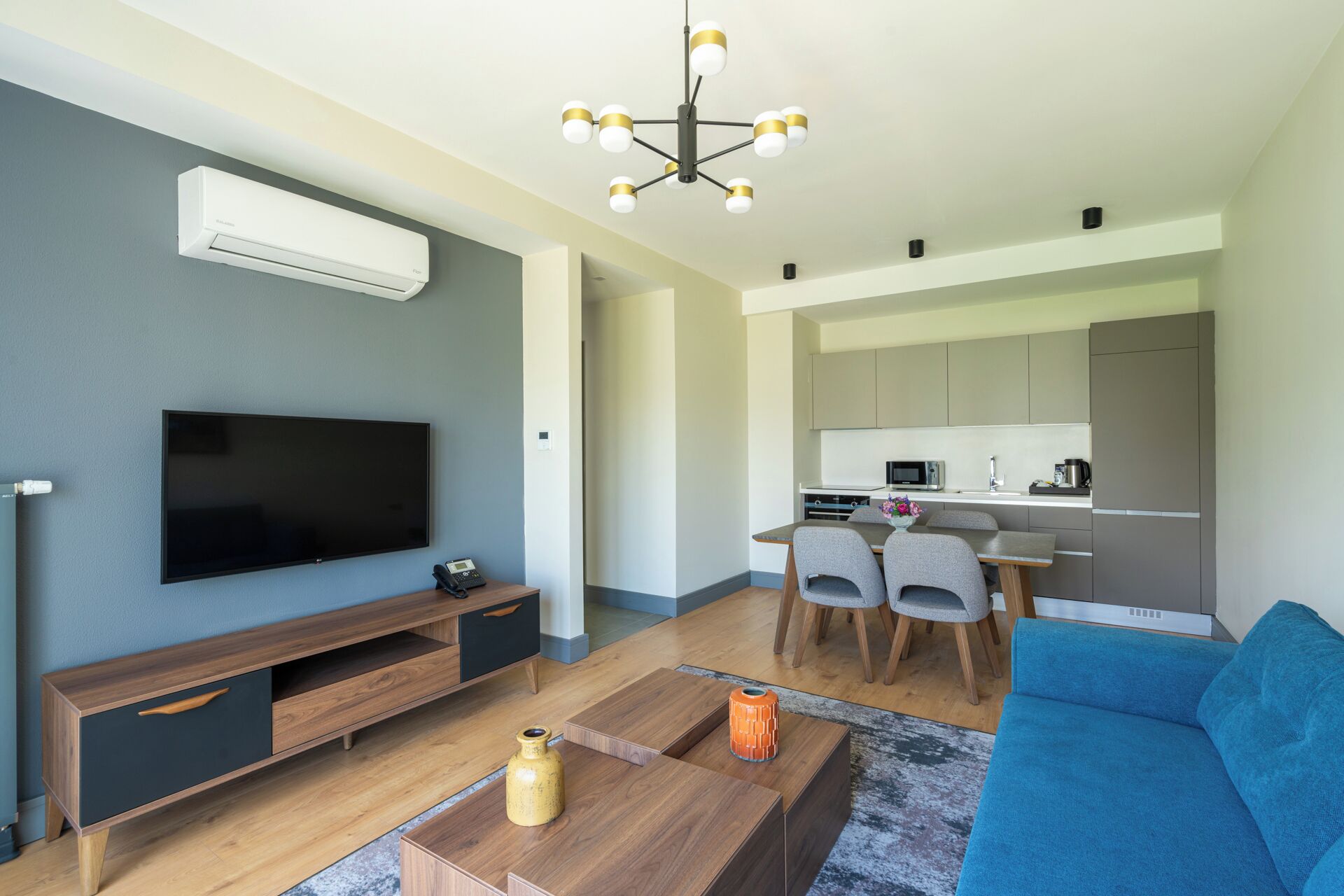 A modern living room with a flat-screen TV, a wooden coffee table, a blue sofa, and a kitchen area with a dining table and chairs.