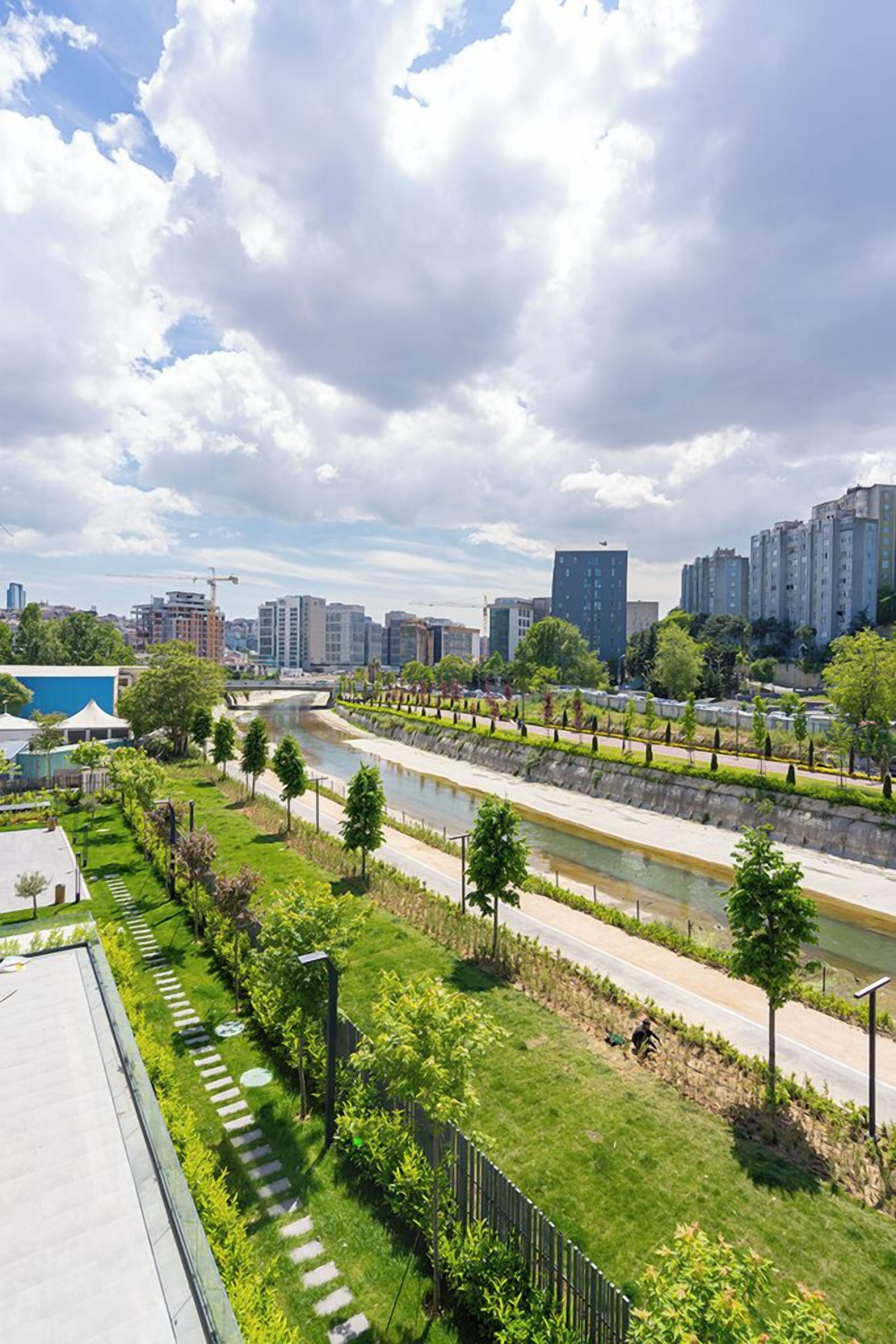 A riverside walkway with greenery, a bridge, and modern buildings in the background.