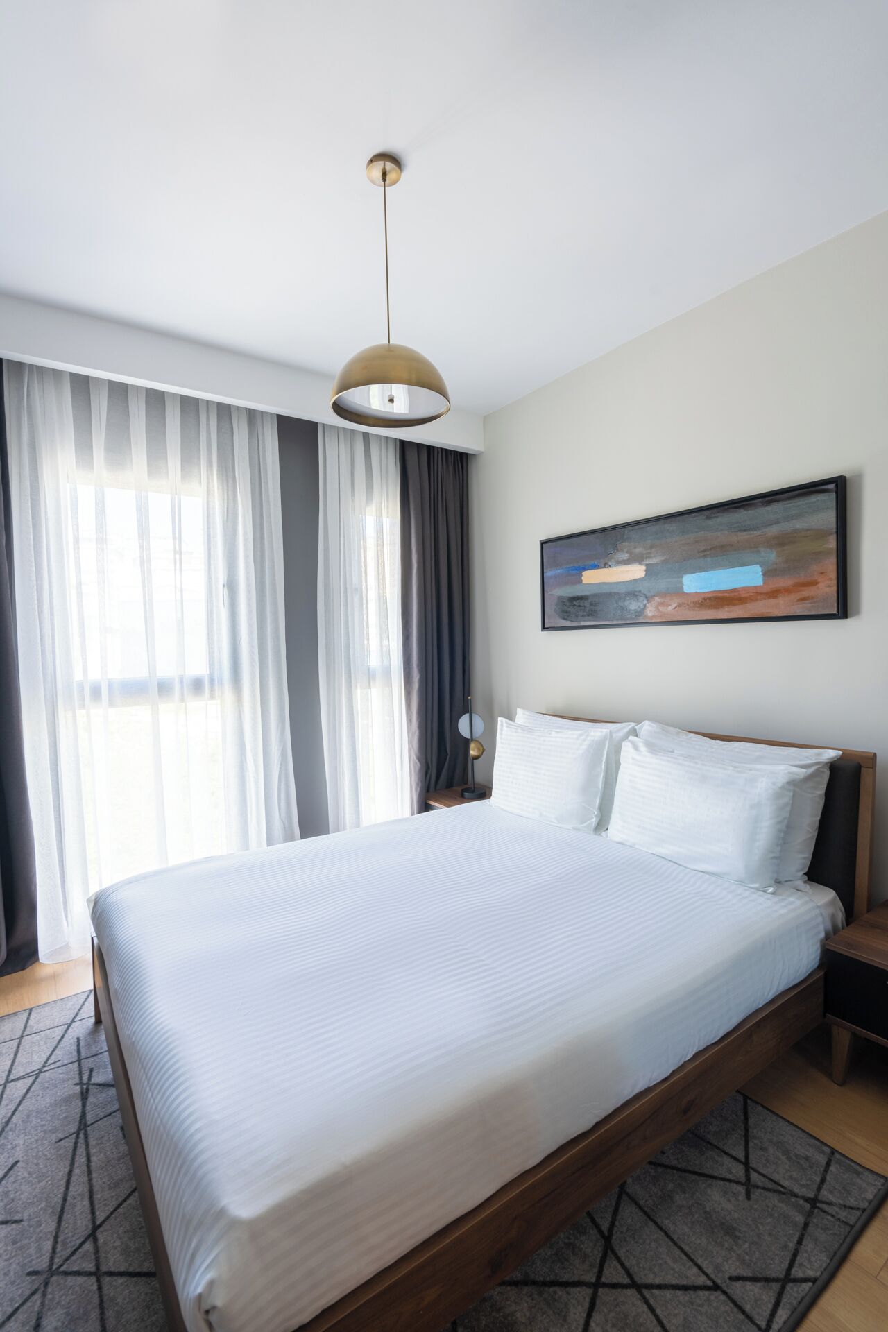 A modern hotel room with a large bed, a round pendant light, a painting on the wall, and a view of the outdoors through sheer curtains.