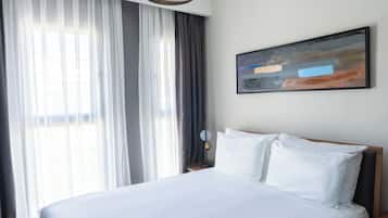 A modern hotel room with a large bed, a round pendant light, a painting on the wall, and a view of the outdoors through sheer curtains.