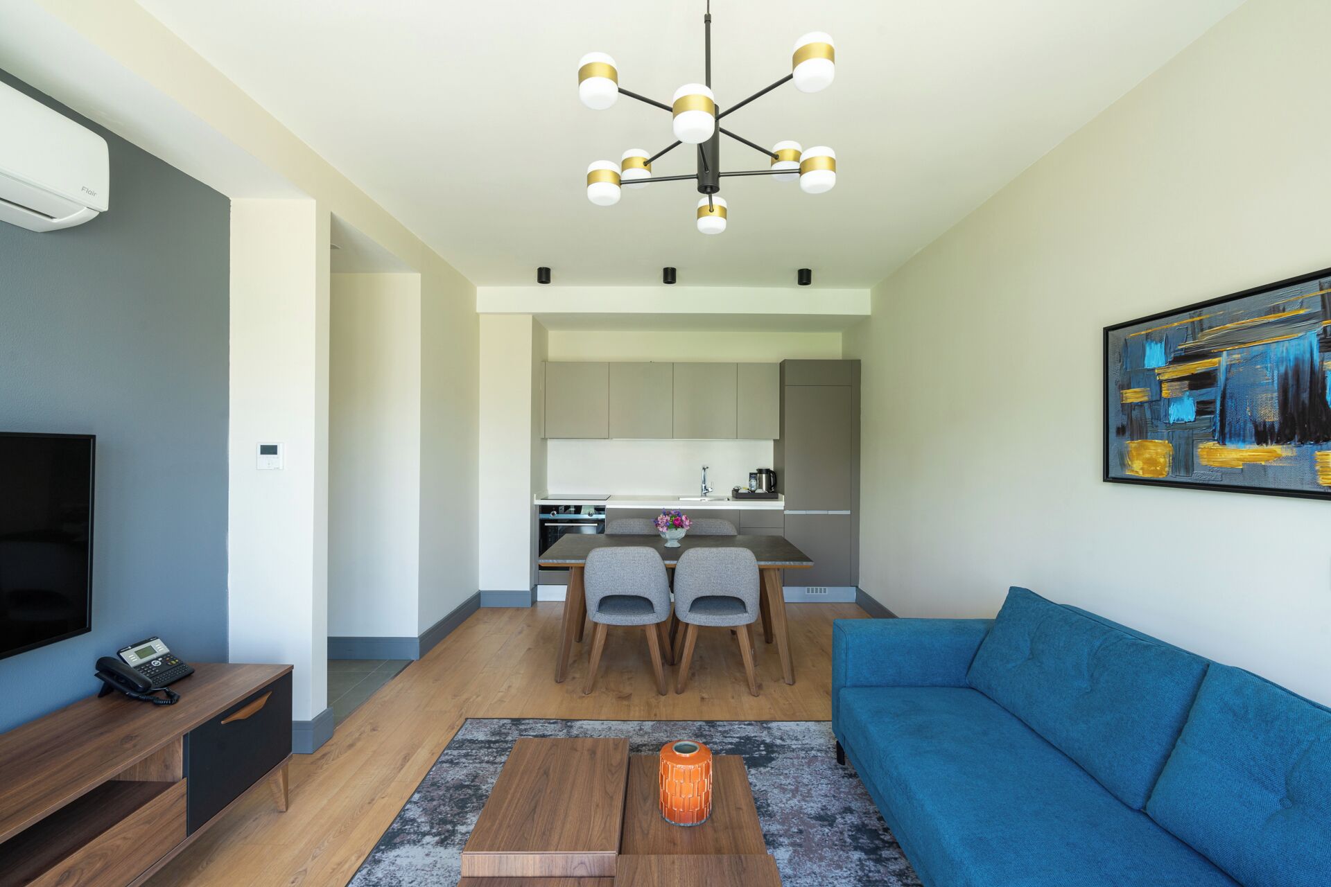 Una sala moderna con un sofá azul, una mesa de centro de madera y un televisor sobre un mueble de madera. Se observa una cocina con una mesa de comedor y sillas, además de un cuadro en la pared.