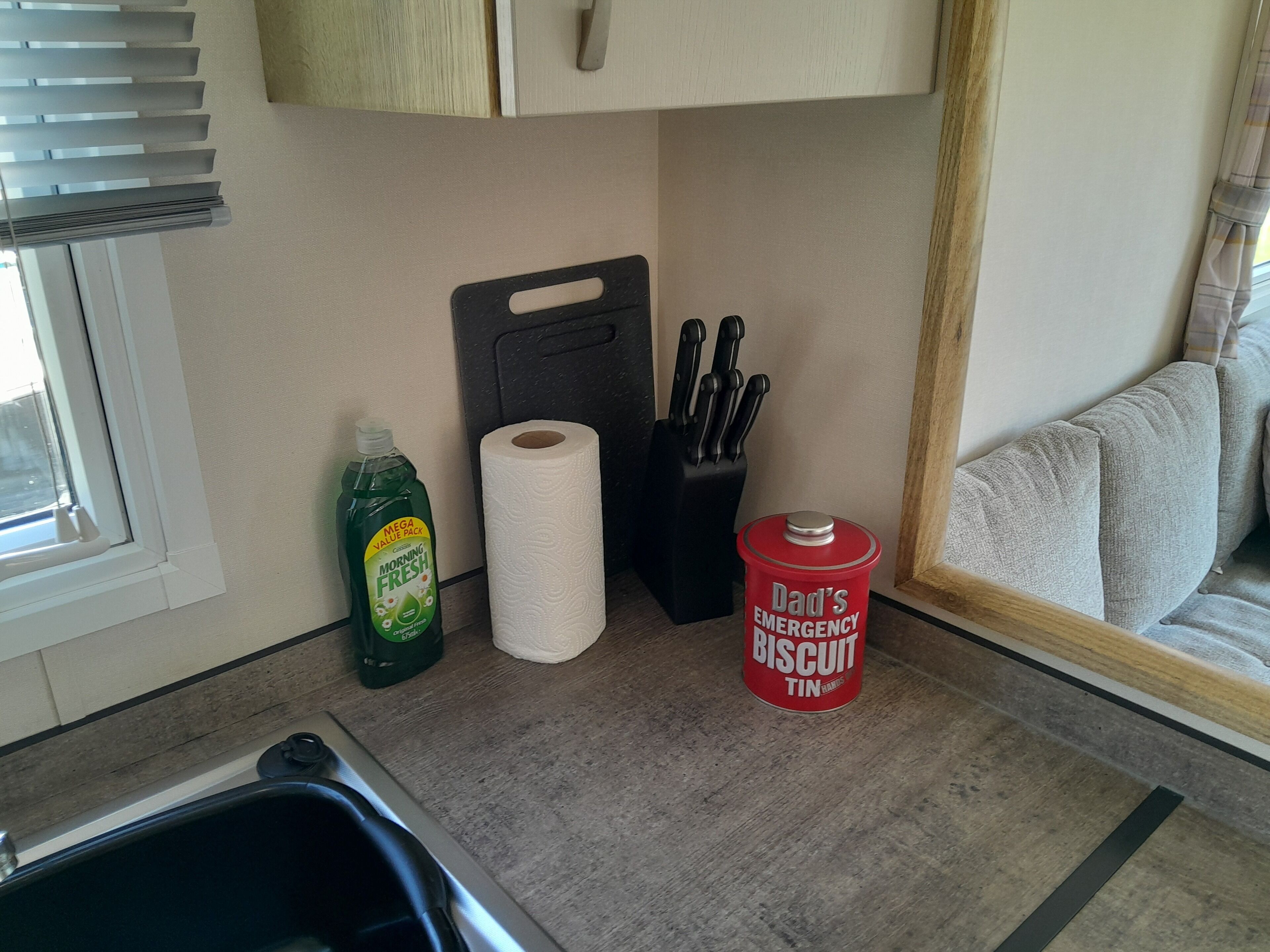 Una encimera de cocina con una botella de detergente, un rollo de toallas de papel, un bloque de cuchillos y una lata con la etiqueta 'La Lata de Galletas de Emergencia de Papá'.