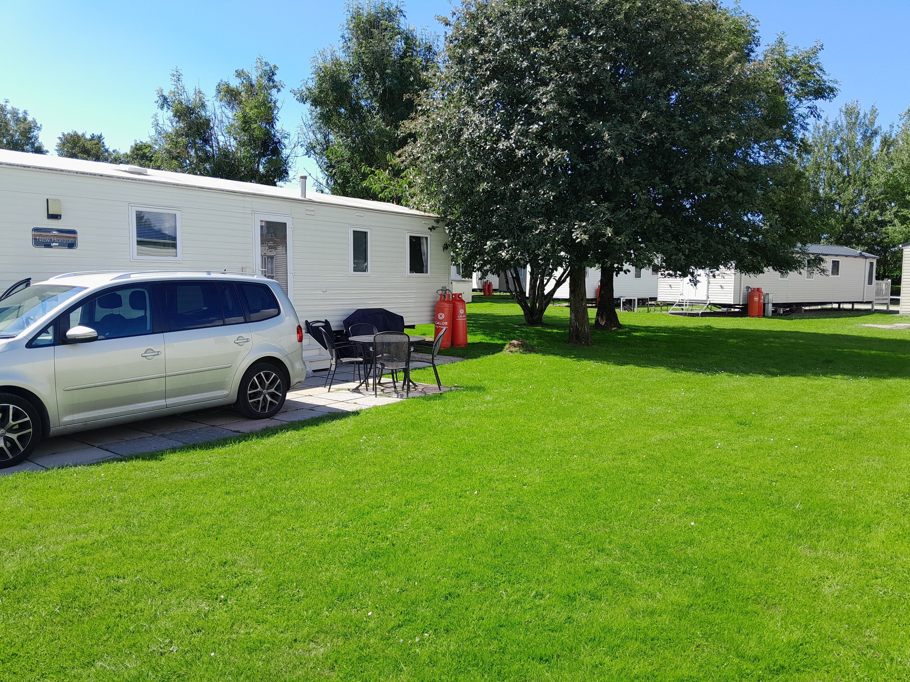 Una caravana blanca estacionada en un césped con una mesa y sillas de patio, rodeada de árboles y otras caravanas.