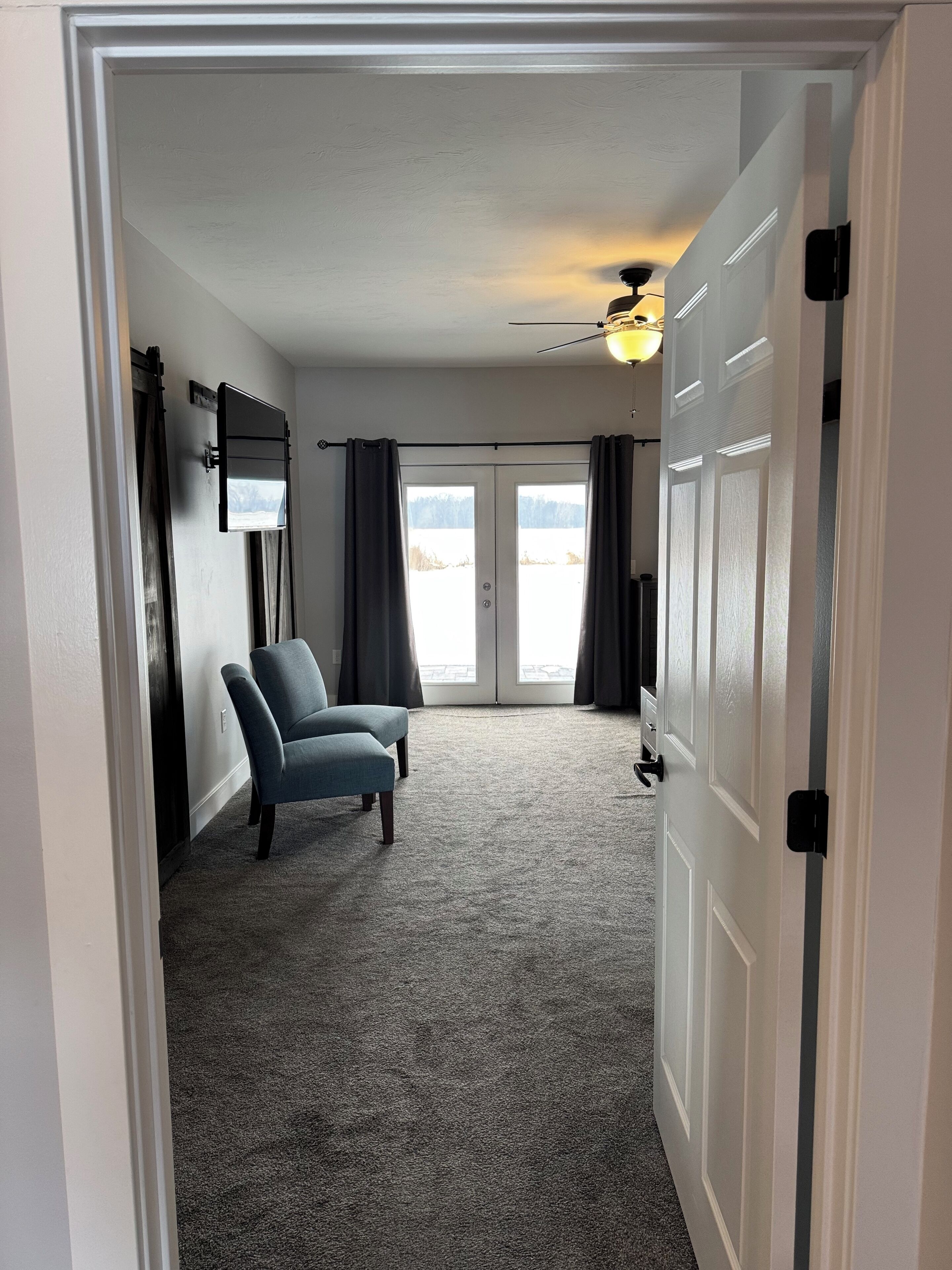 A room with a grey carpet, a blue chair, a ceiling fan, and a door with a view to the outside.