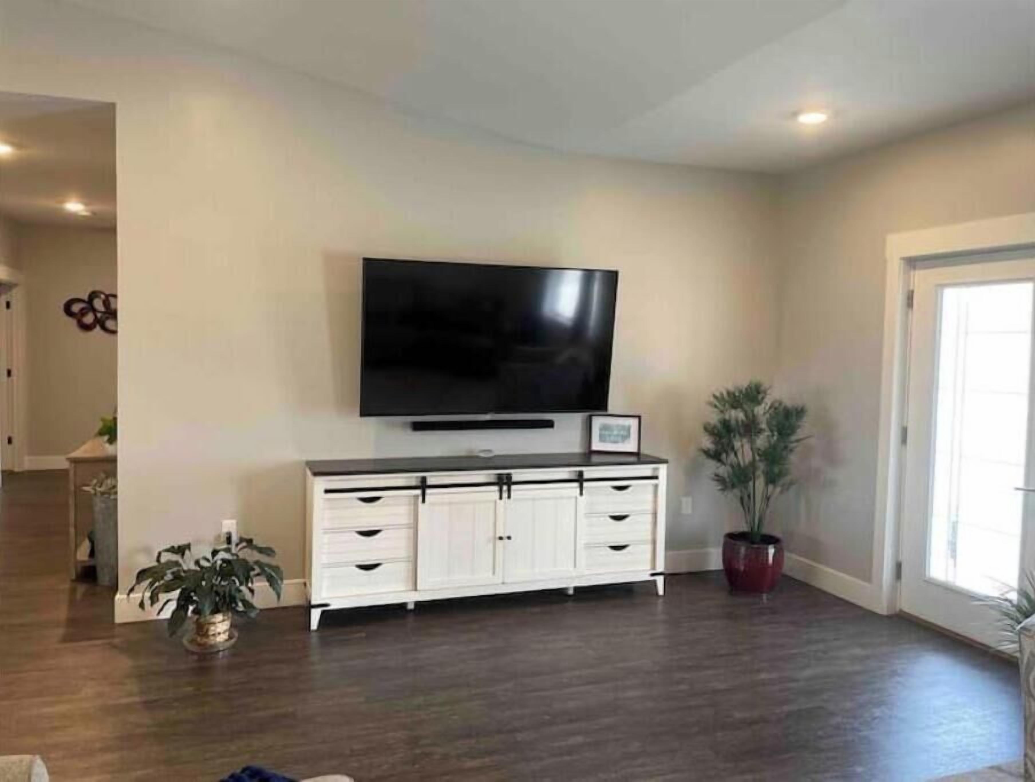 Een ruime woonkamer met een flatscreen televisie bevestigd aan een witte kast, donker houten vloeren en een pot met een plant.