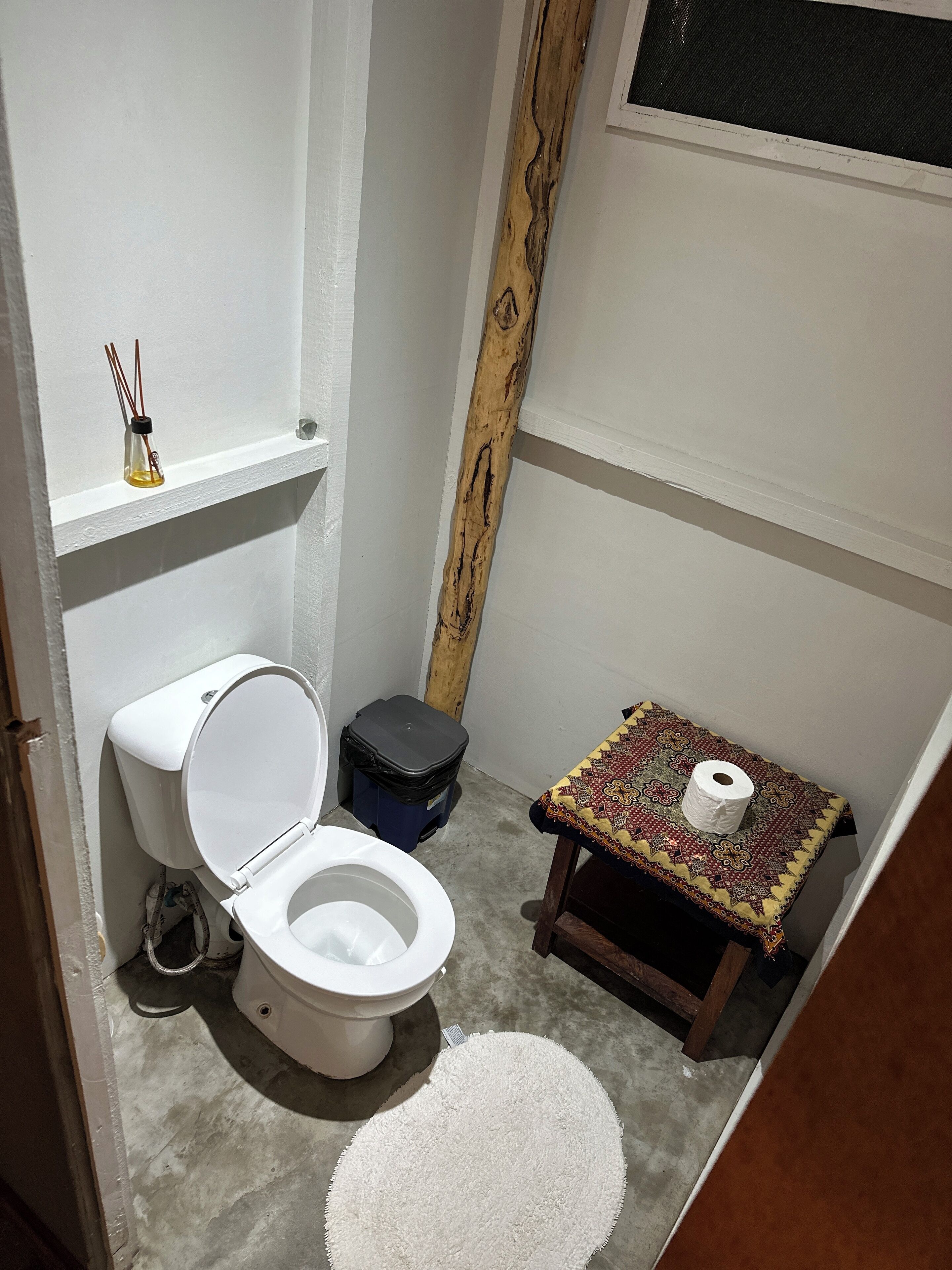 Een kleine, compacte badkamer met een wit toilet, een klein prullenbakje, een houten krukje met een tafelkleed met patroon, en een witte vloerkleed.