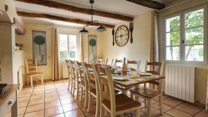Un coin repas avec une table en bois dressée pour quatre personnes, entourée de chaises. Il y a une fenêtre avec des rideaux, une horloge au mur et un radiateur sous la table.