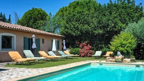 Une piscine entourée de chaises longues et de parasols, elle-même bordée de verdure et d’un bâtiment doté d’un toit en tuiles.