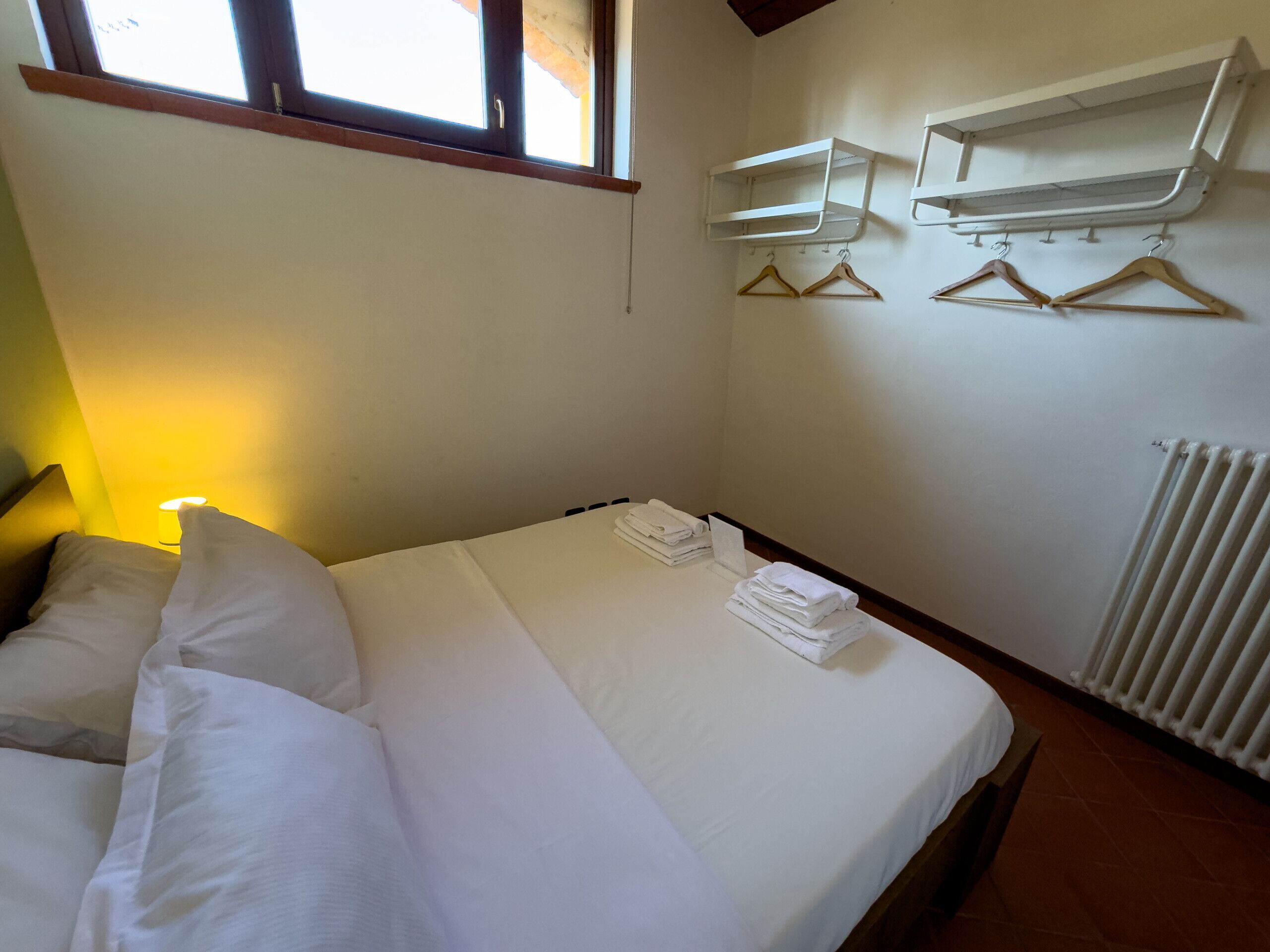 A neatly made bed with white linens, a bedside lamp, and a radiator. There are two wall-mounted shelves above a radiator, and a window with a wooden frame is visible.