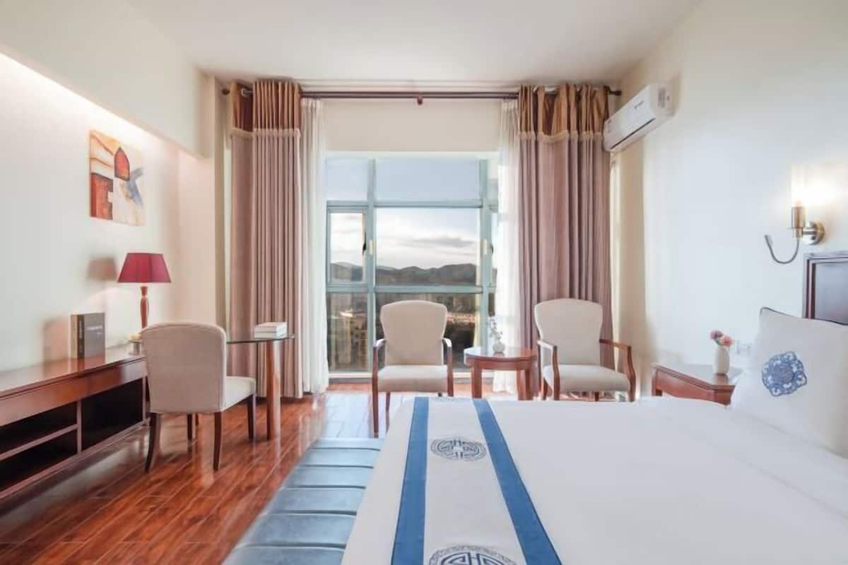Habitación de hotel con una cama grande, dos sillones, un escritorio y vistas amplias por un ventanal.