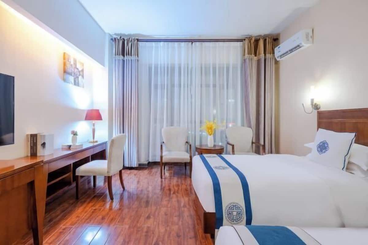 Una habitación de hotel con piso de madera, una cama con cubrecama azul y blanco, un escritorio con lámpara, una silla y un televisor.