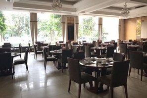 Un restaurant amb taules i cadires rodones, grans finestres amb cortines i un penjallum.