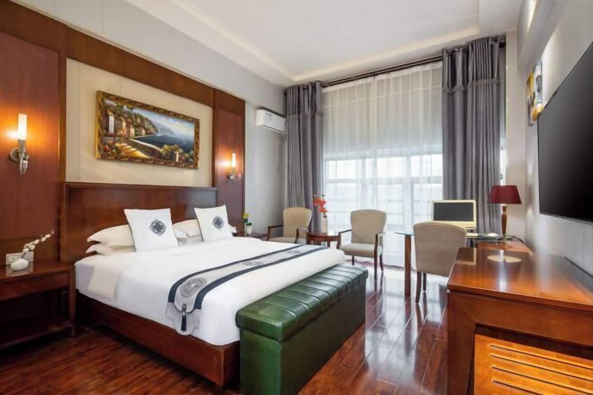 Habitación de hotel con una cama grande, un banco tapizado de verde, un escritorio con silla, un televisor y un cuadro en la pared.