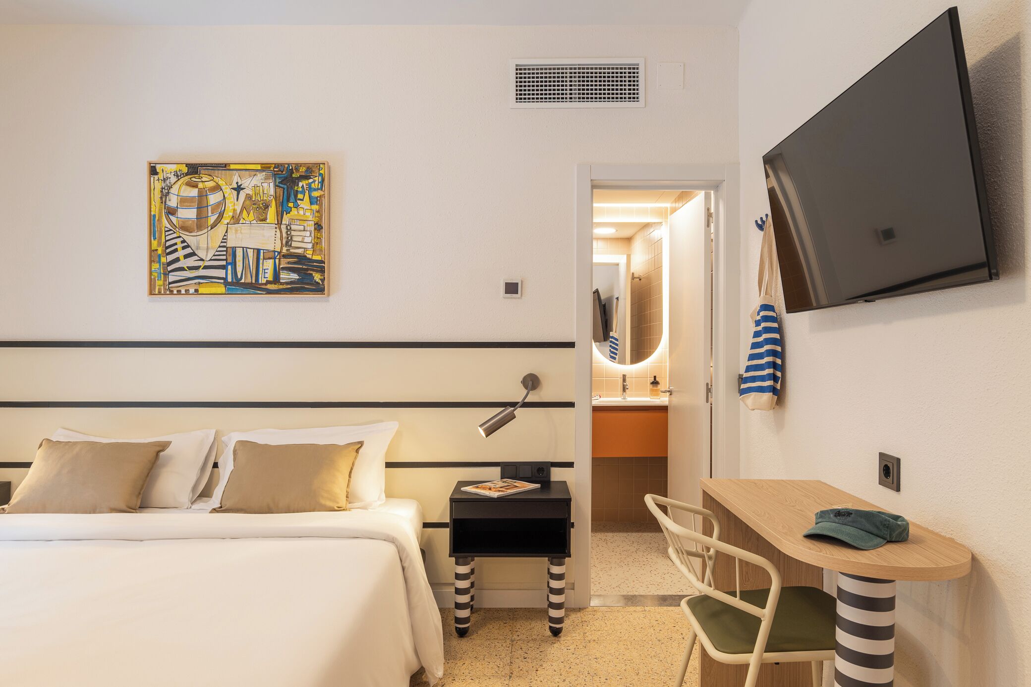 A hotel room with a bed, bedside table, chair, TV, artwork, and a bathroom visible through an open door.