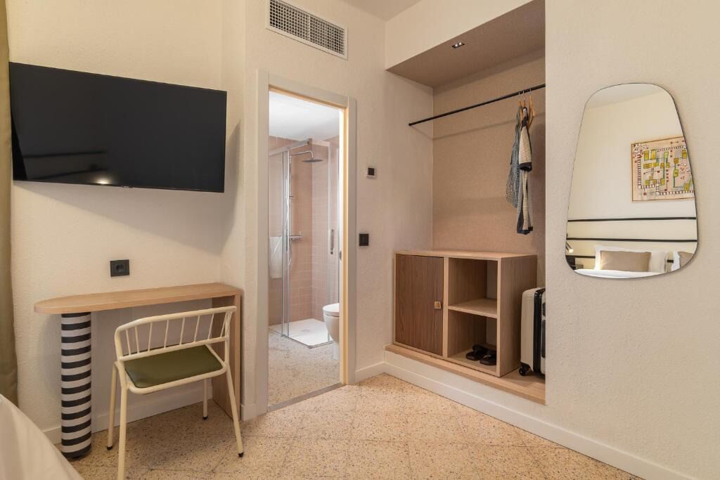 A compact hotel room with a bed, a small desk with a chair, a shower area, and a wardrobe with a mirror.