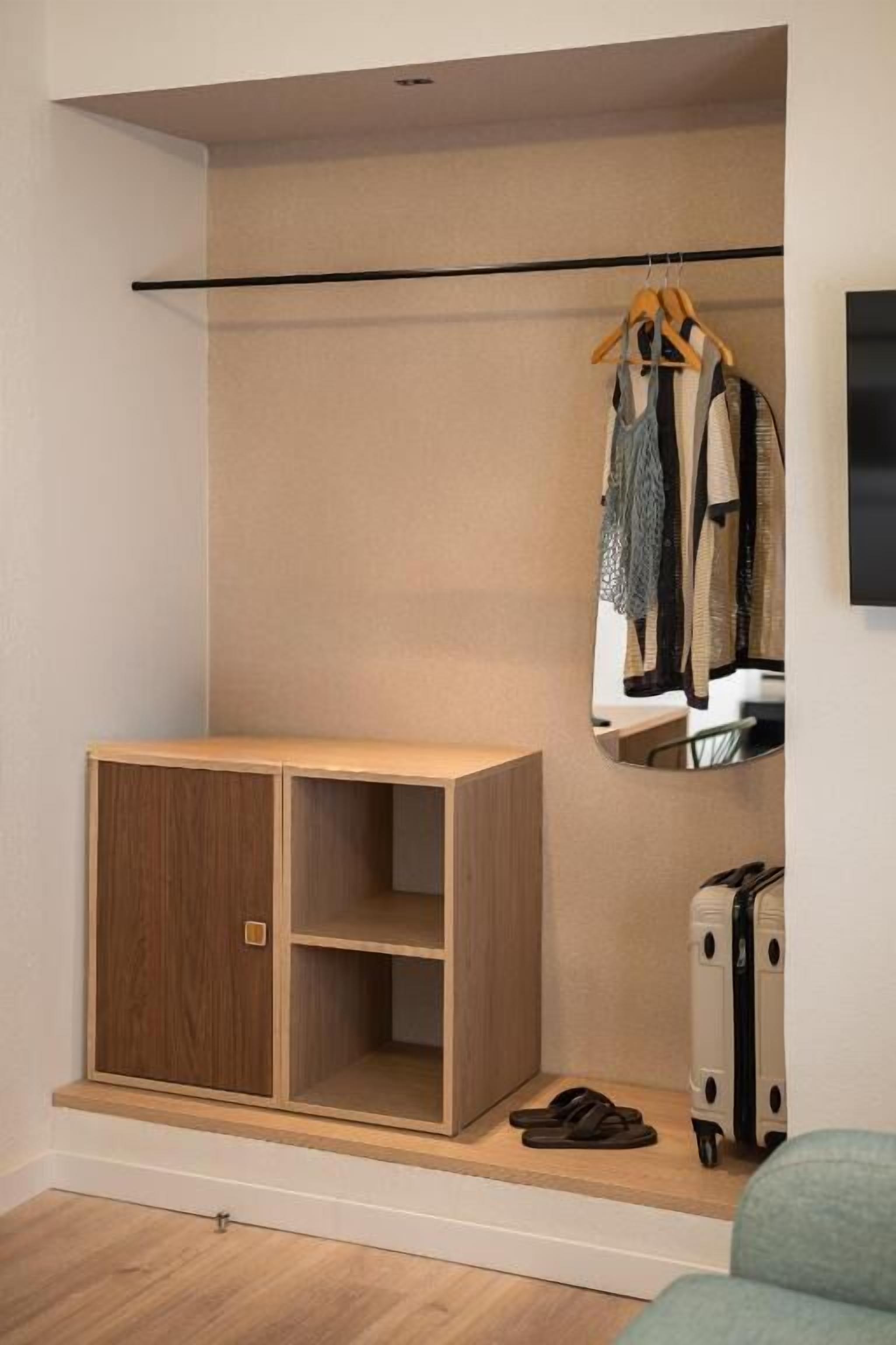 Una habitación con un armario de madera, un perchero con ropa, una maleta y un espejo en la pared.