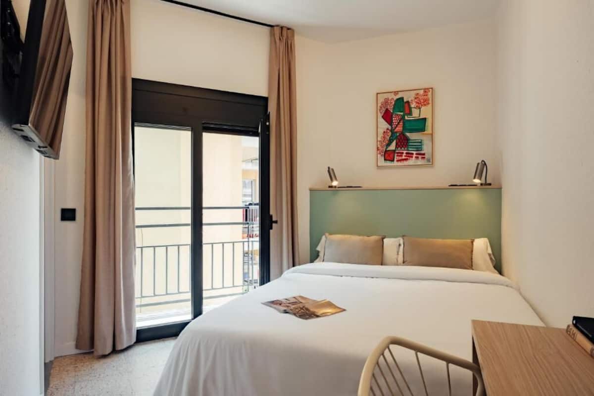 A hotel room with a bed, a desk, a chair, a window with curtains, and a painting on the wall.