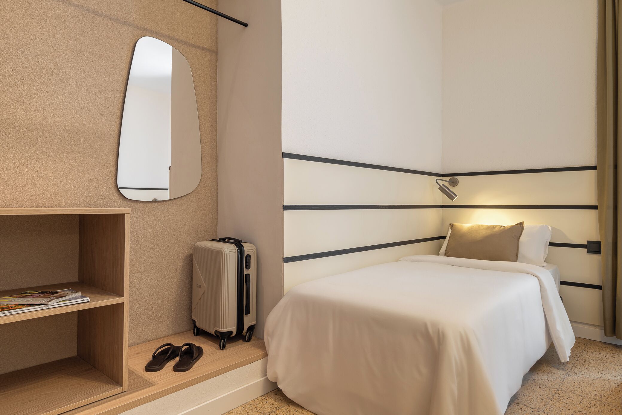 A hotel room with a bed, a desk with a mirror, a suitcase, and a pair of shoes.