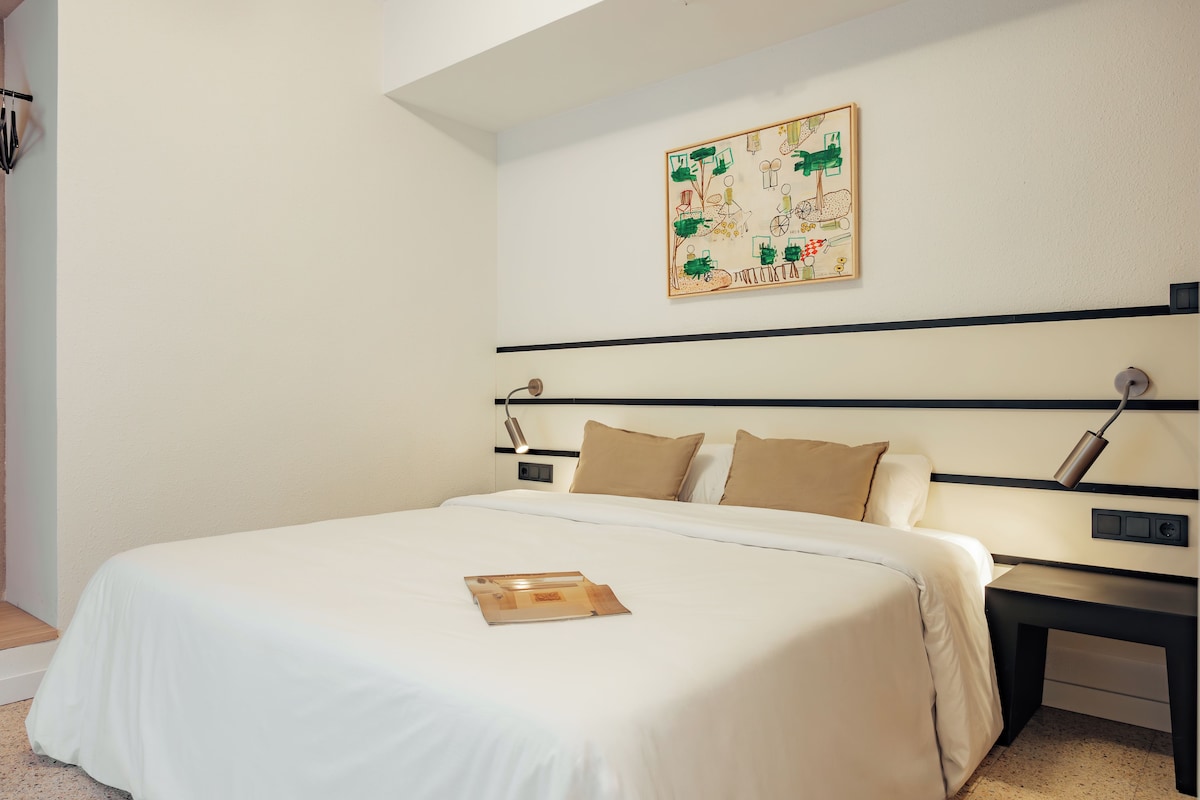 A neatly made bed with a white bedspread and a book on it, a bedside table with a lamp, and a wall-mounted artwork above the bed.