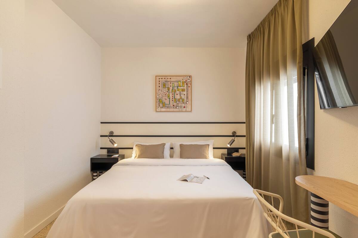 A hotel room with a neatly made bed, a bedside table with a lamp, a small table, and a large window with curtains.