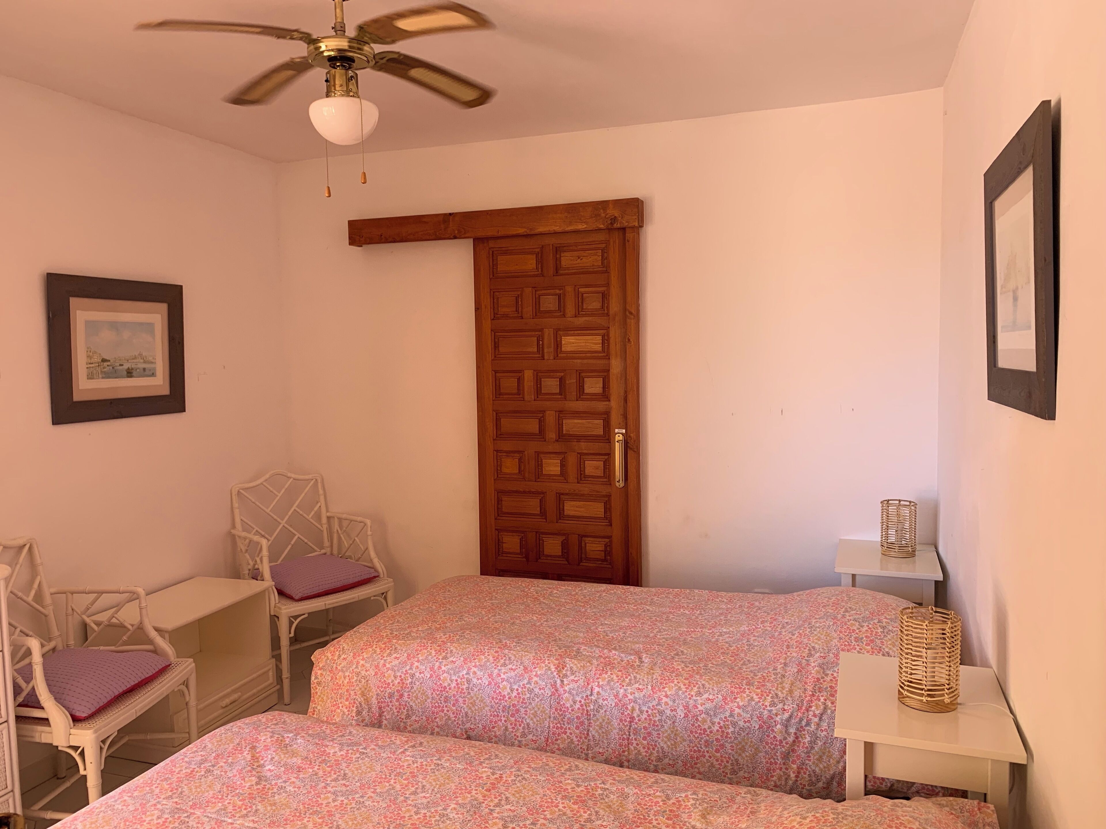 Ein Schlafzimmer mit zwei Betten, einer Holztür, einem Deckenventilator, einem Stuhl, einem kleinen Tisch und einem gerahmten Bild an der Wand.