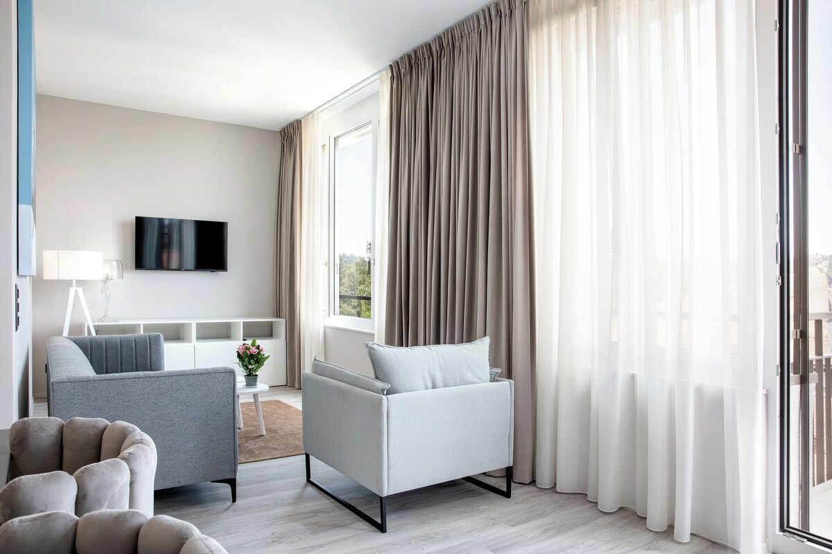 Una sala de estar moderna con un sofá gris, un sillón más pequeño, un televisor de pantalla plana y un amplio ventanal con cortinas.
